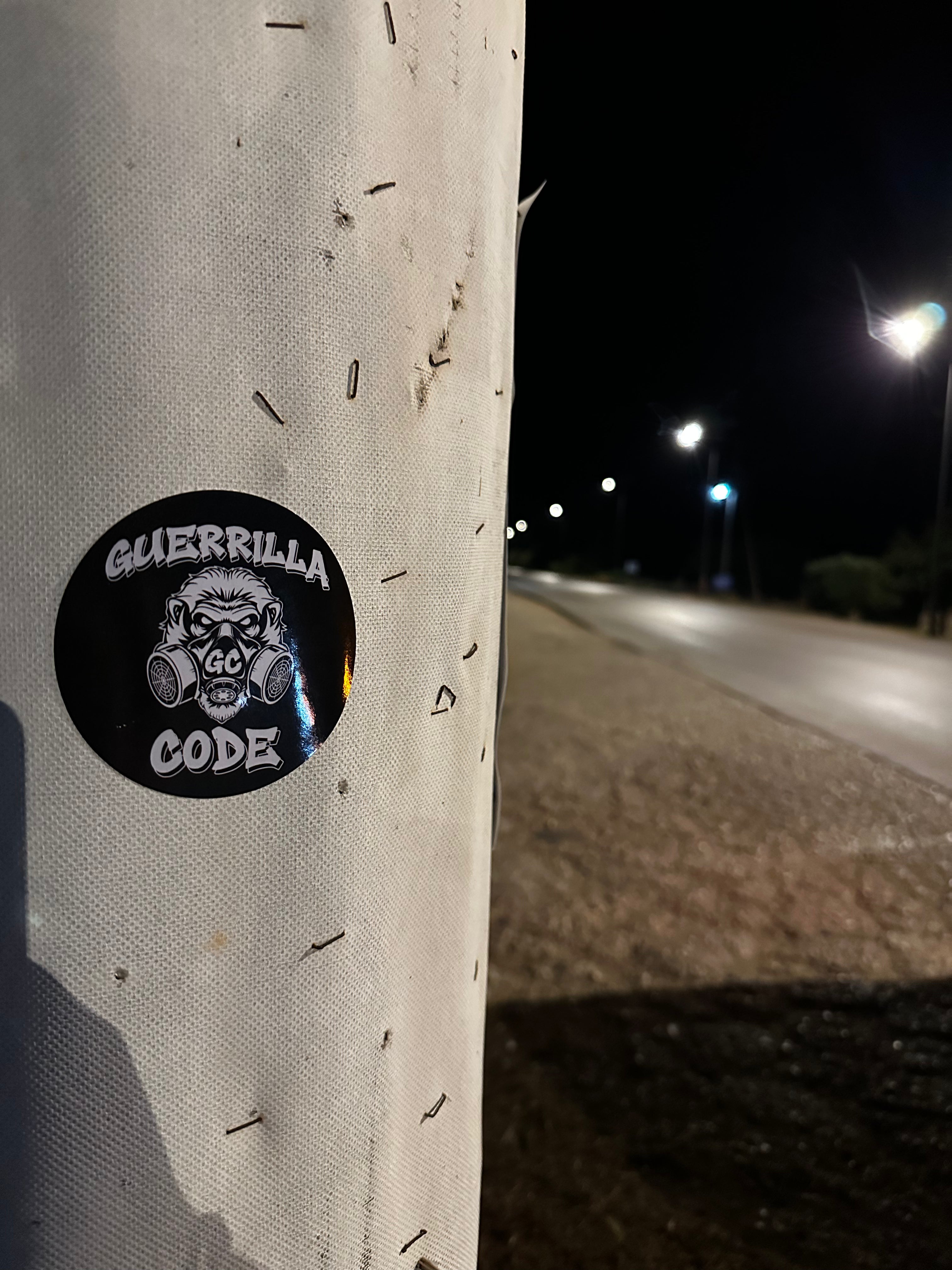 A round black sticker with a white gorilla wearing a gas mask and the words 'GUERRILLA CODE' is shown on a white pole at night, with a blurred road and streetlights in the background.